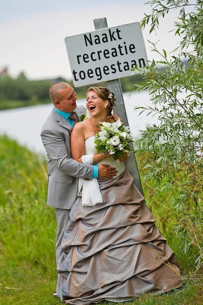 De bruid leunt lachend tegen de bruidegom naast een bordje ‘Naaktrecreatie toegestaan’ aan het water