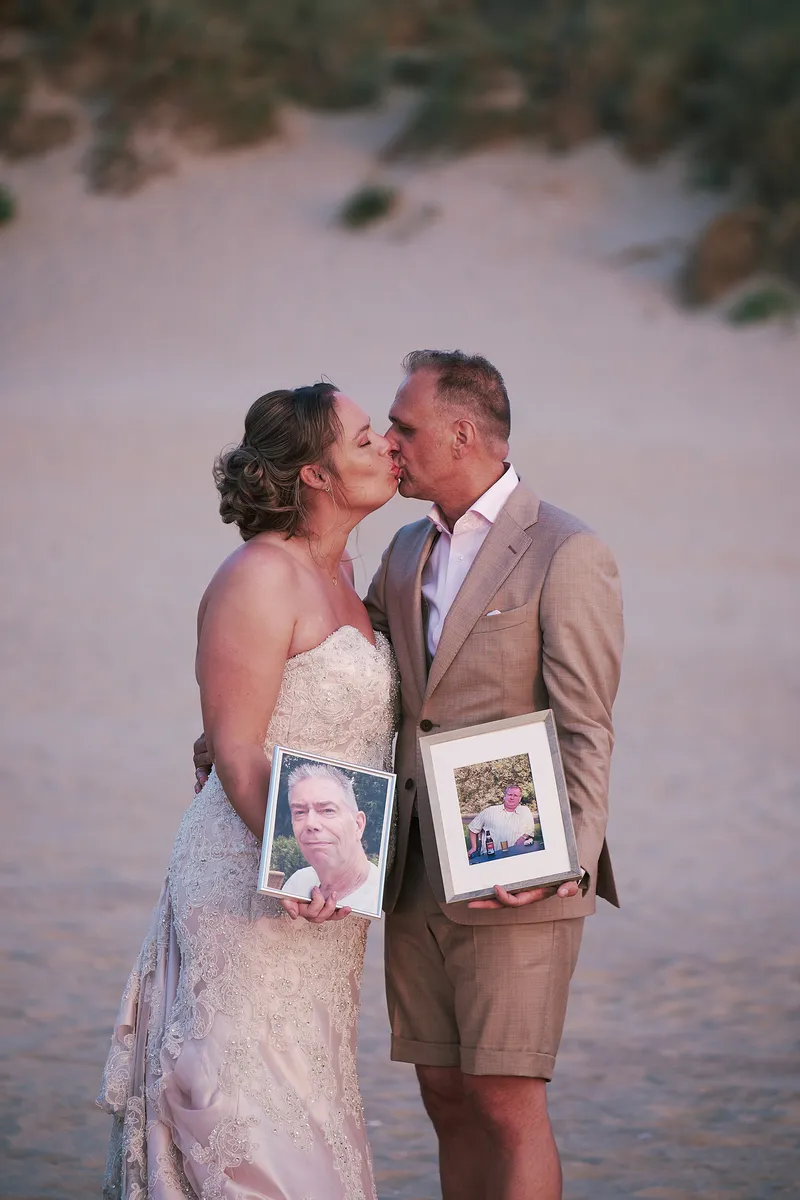 Mirjam en Jorg kussen elkaar in de duinen, met de foto’s van hun vaders in hun handen